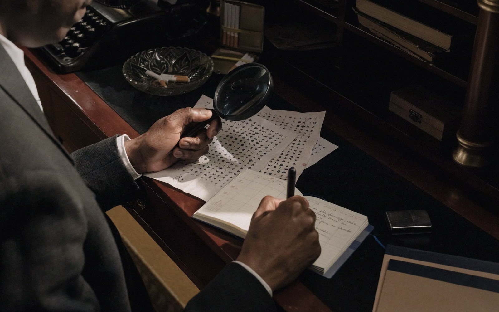A man examines coded symbol charts with a magnifying glass while writing in a notebook at a desk