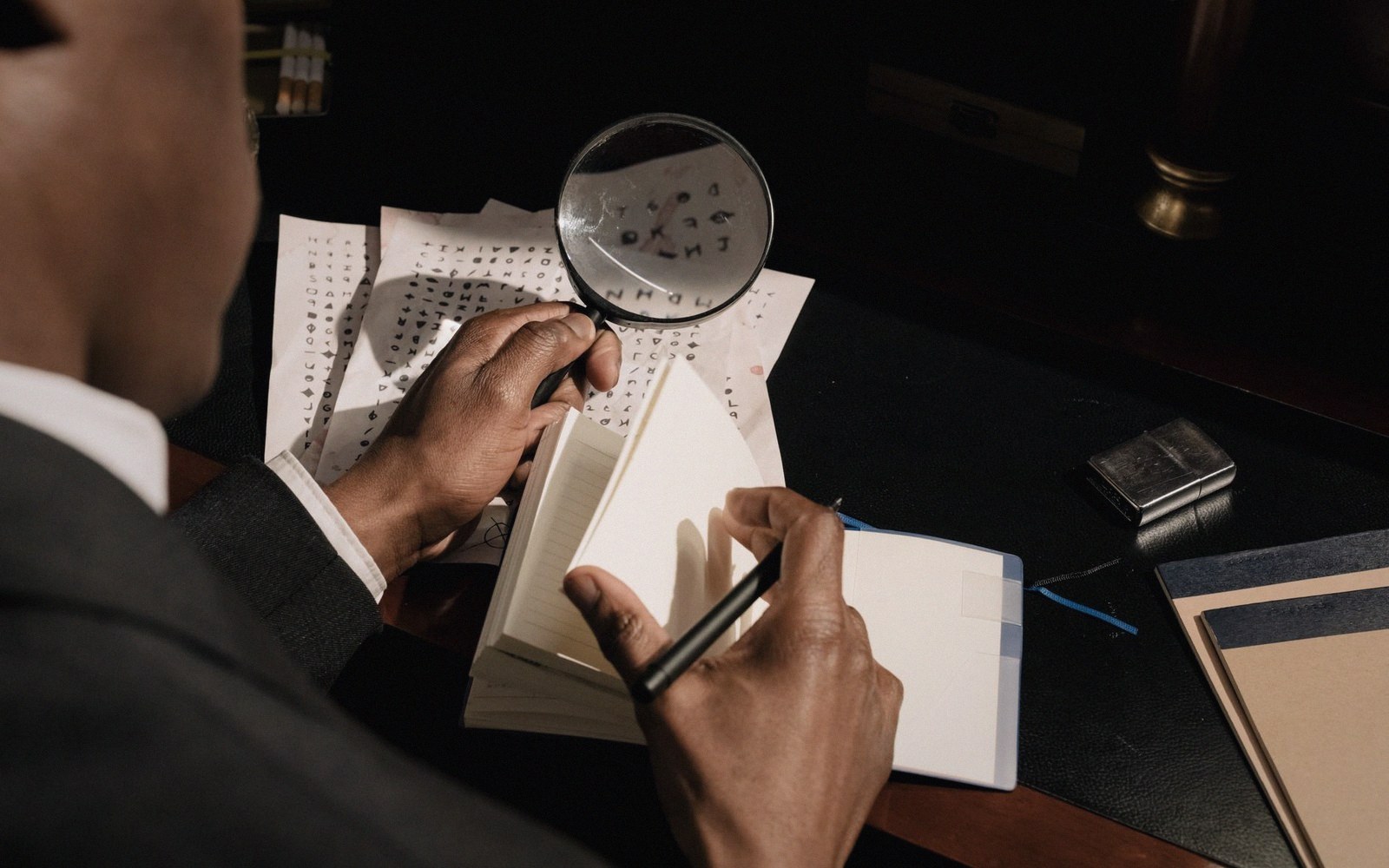 A man uses a magnifying glass to examine coded pages while taking notes at a desk