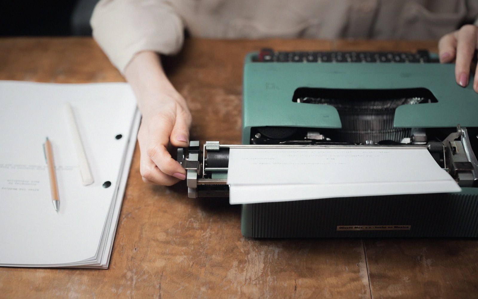 Hands feeding paper into a vintage teal typewriter beside a stack of typed pages