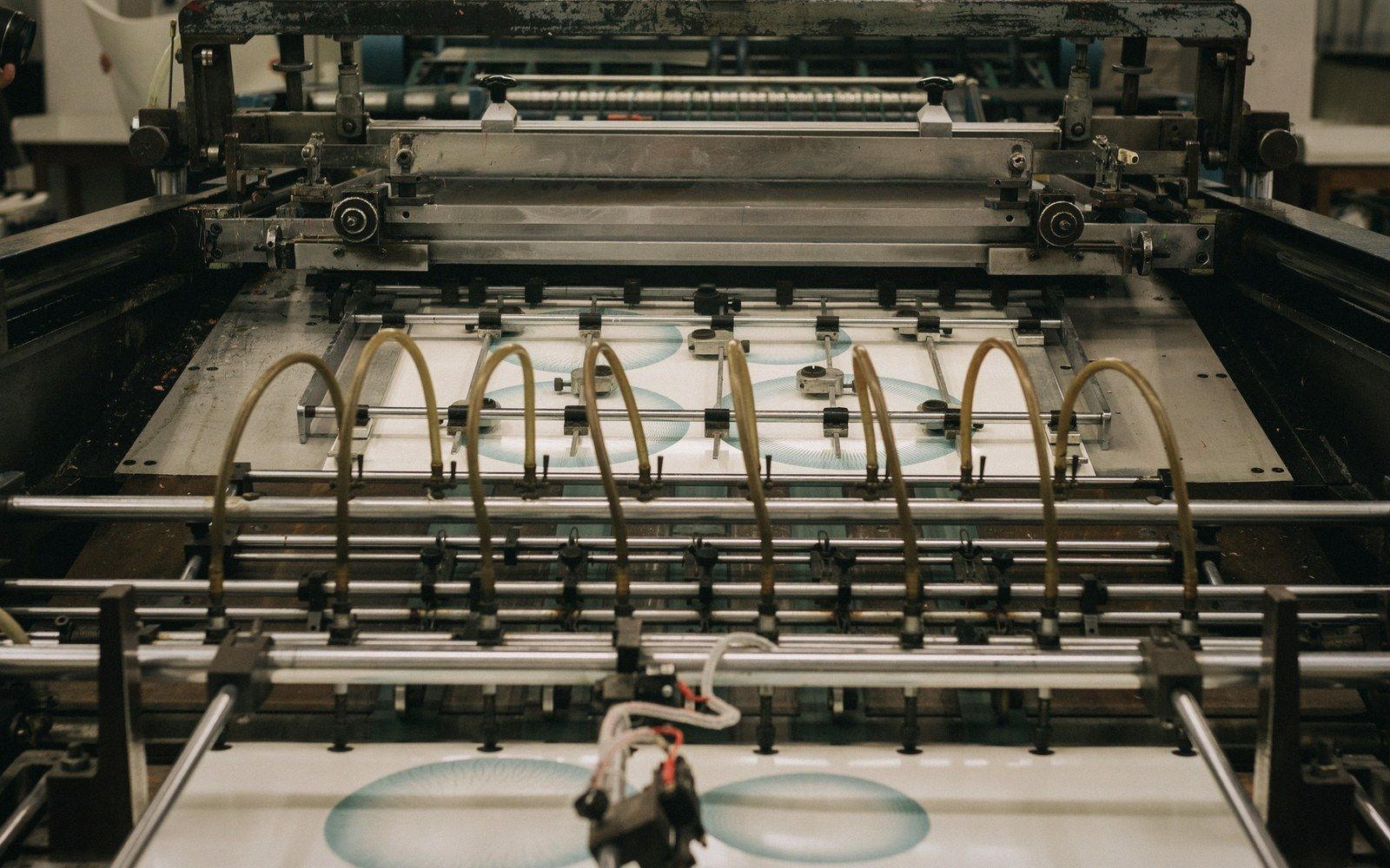 Industrial printing press feeding paper sheets with circular teal patterns through metal rollers